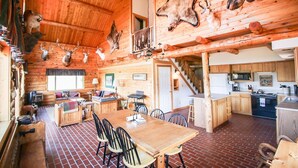 Interno di un rifugio rustico con zona pranzo, angolo cottura e soffitto decorato con trofei di animali appesi.