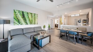 A modern living room with a gray sectional sofa, a glass coffee table, and a dining area with blue upholstered chairs. - Water Park + Pool | Shuttle to Disney World (Orlando)