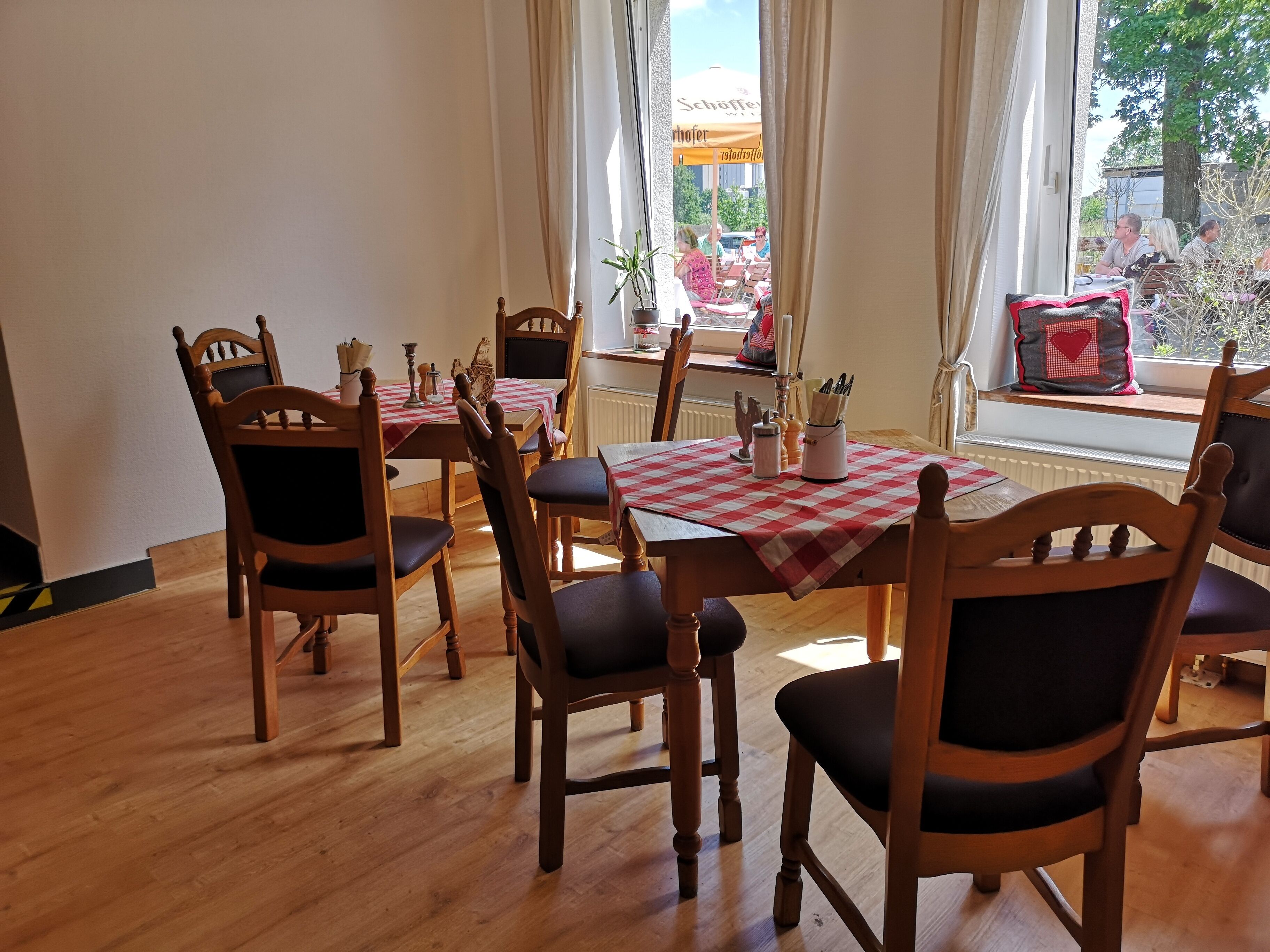Un comedor con sillas y mesas de madera, vistas al exterior a través de grandes ventanales y un mantel a cuadros rojo y blanco.