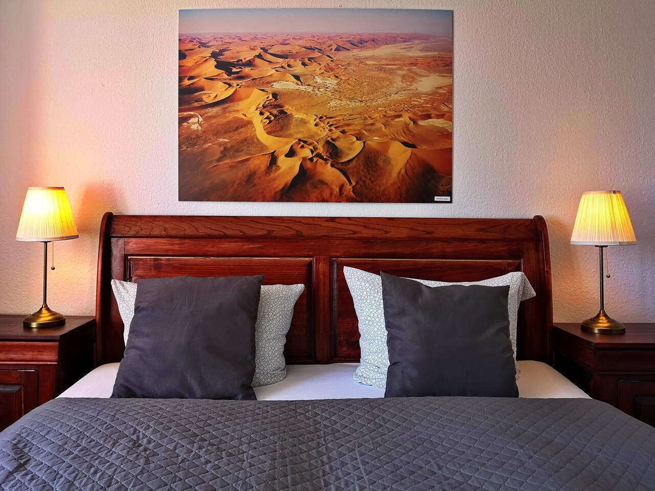 Ein Hotelzimmer mit einem hölzernen Kopfteil, zwei Nachttischlampen, einem großen Gemälde von einer Wüstenlandschaft an der Wand und einem Bett mit einer gesteppten Bettdecke.