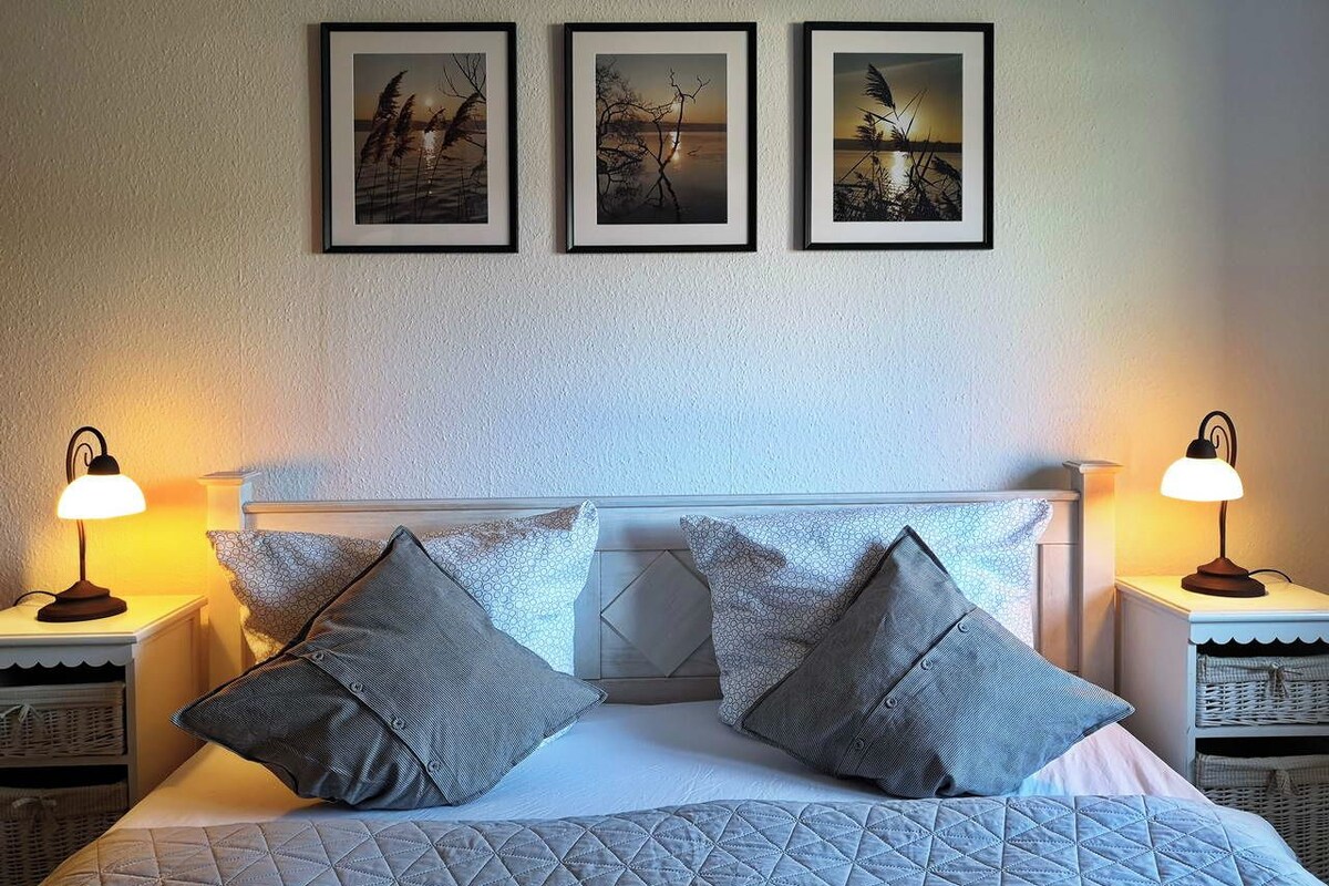 Una cama bien hecha con cubrecolchón acolchado, dos mesitas de noche con lámparas, y tres cuadros enmarcados en la pared.