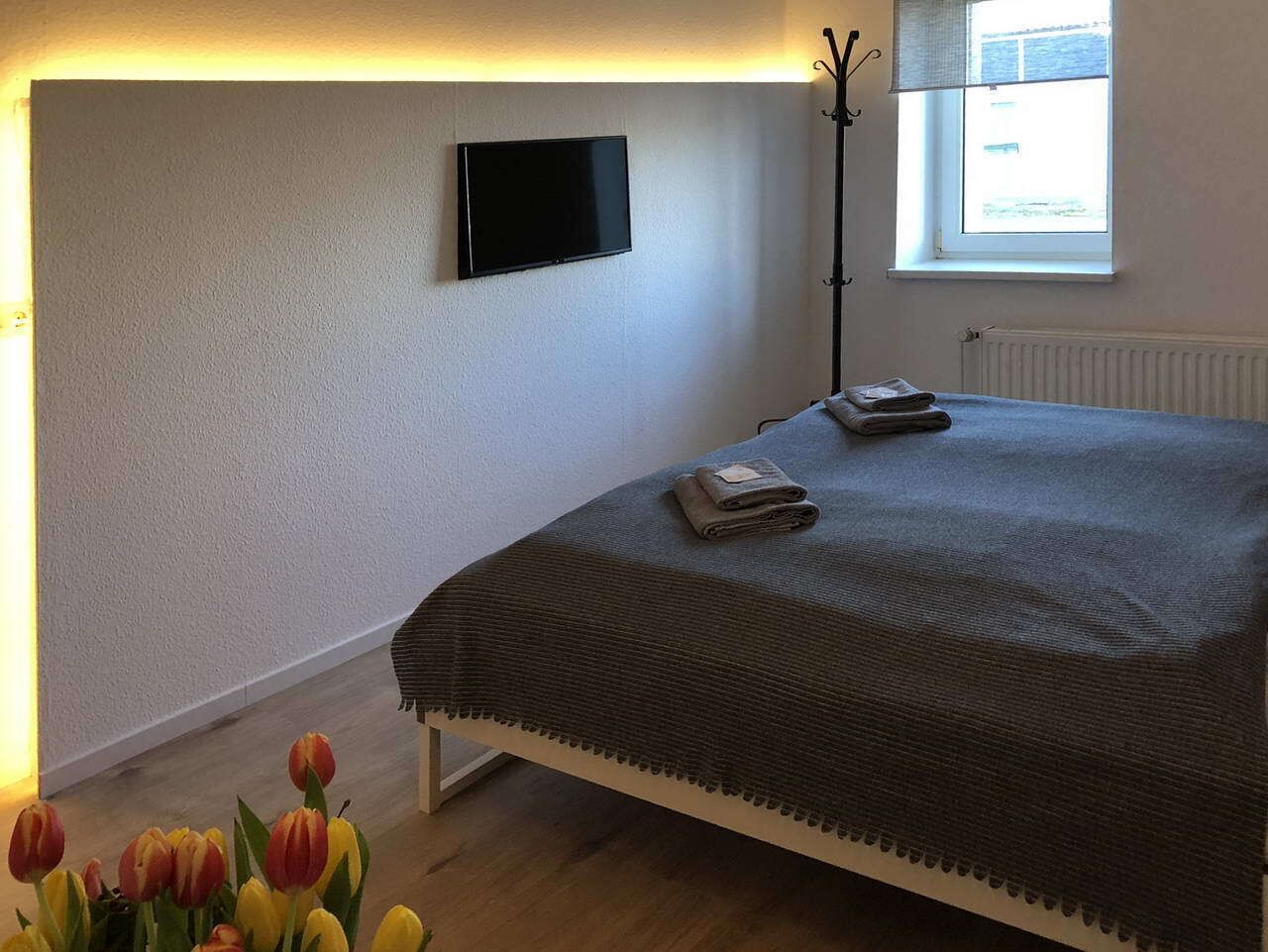 A bedroom with a bed, a flat-screen TV mounted on the wall, a window with a view, and a floor lamp.