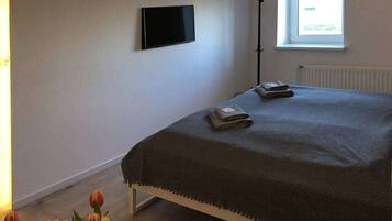 A bedroom with a bed, a flat-screen TV mounted on the wall, a window with a view, and a floor lamp.