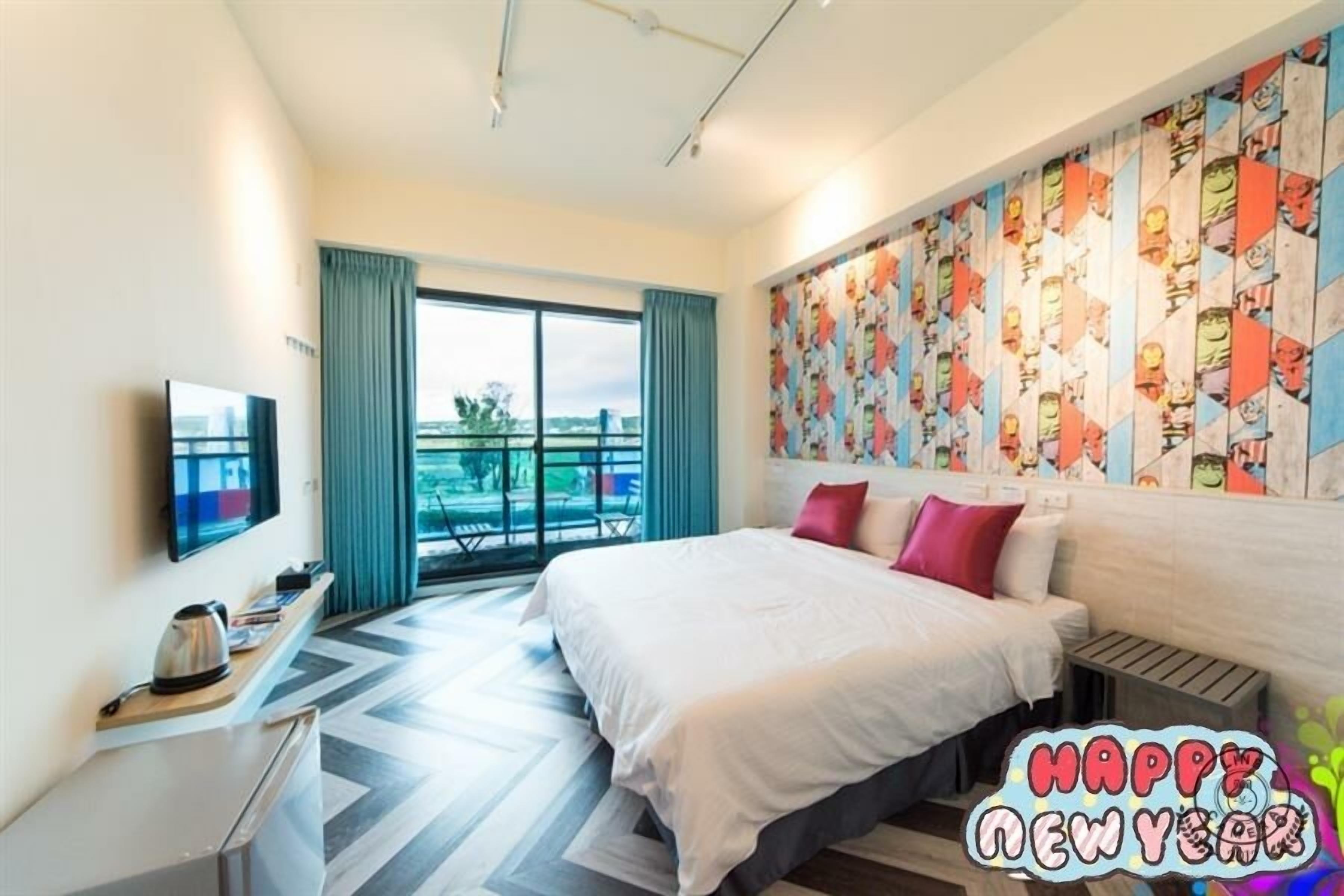 A hotel room with a bed, a television, a kettle, a window with curtains, and a colorful wall mural.