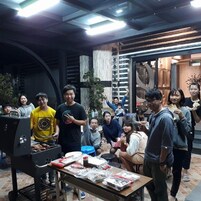 Un grupo de personas reunidas alrededor de una parrilla en una habitación con diseño moderno e industrial, que incluye vigas a la vista y una variedad de asientos.