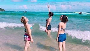 Três pessoas em águas rasas numa praia, com céu limpo e barcos ao longe.