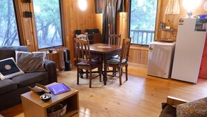 Interior acogedor de una cabaña con una mesa de comedor de madera, sillas, un sofá, una pequeña zona de cocina con refrigerador y un gran ventanal con vistas al bosque.