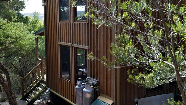 Una cabaña de madera con techo de metal, una escalera que lleva a una terraza y dos tanques de propano grandes en el exterior.