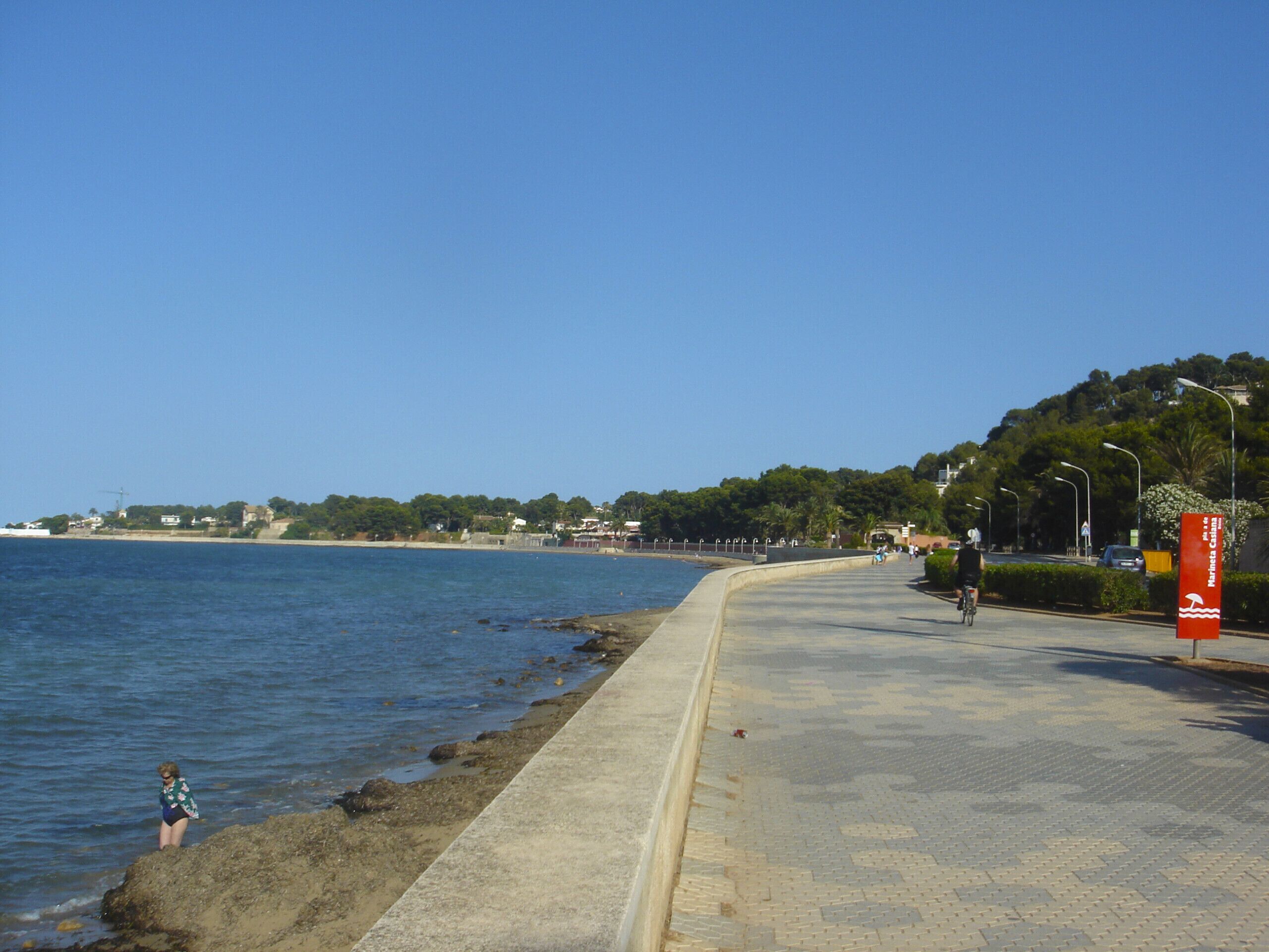 A coastal walkway with a paved path, a cyclist, and a child near the water.