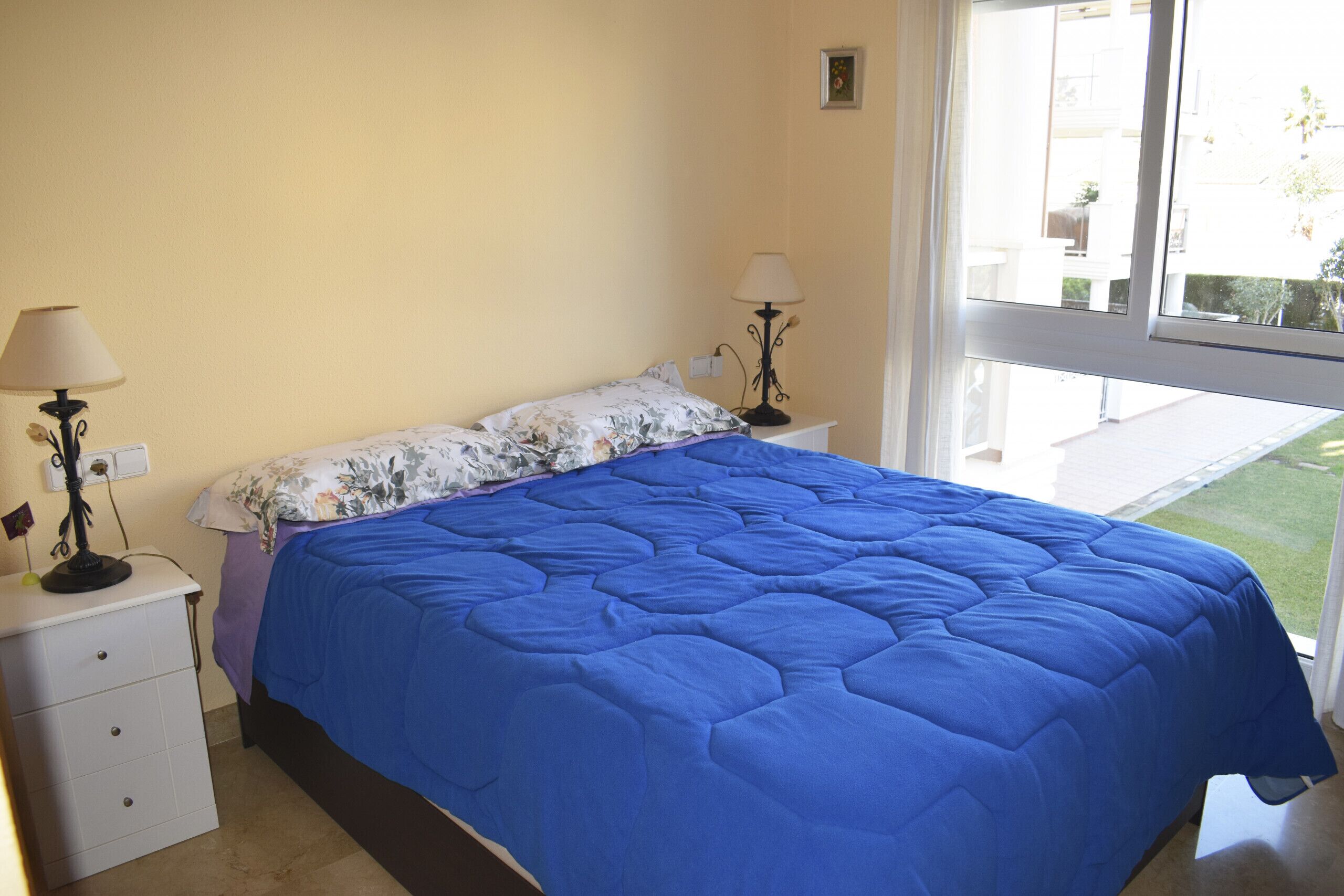 A bedroom with a large bed, two bedside tables with lamps, a window with a view of the outdoors, and a small wall-mounted light.