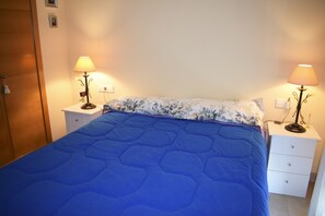 A bedroom with a blue quilted bedspread, two bedside tables with lamps, a wooden wardrobe, and a white nightstand.
