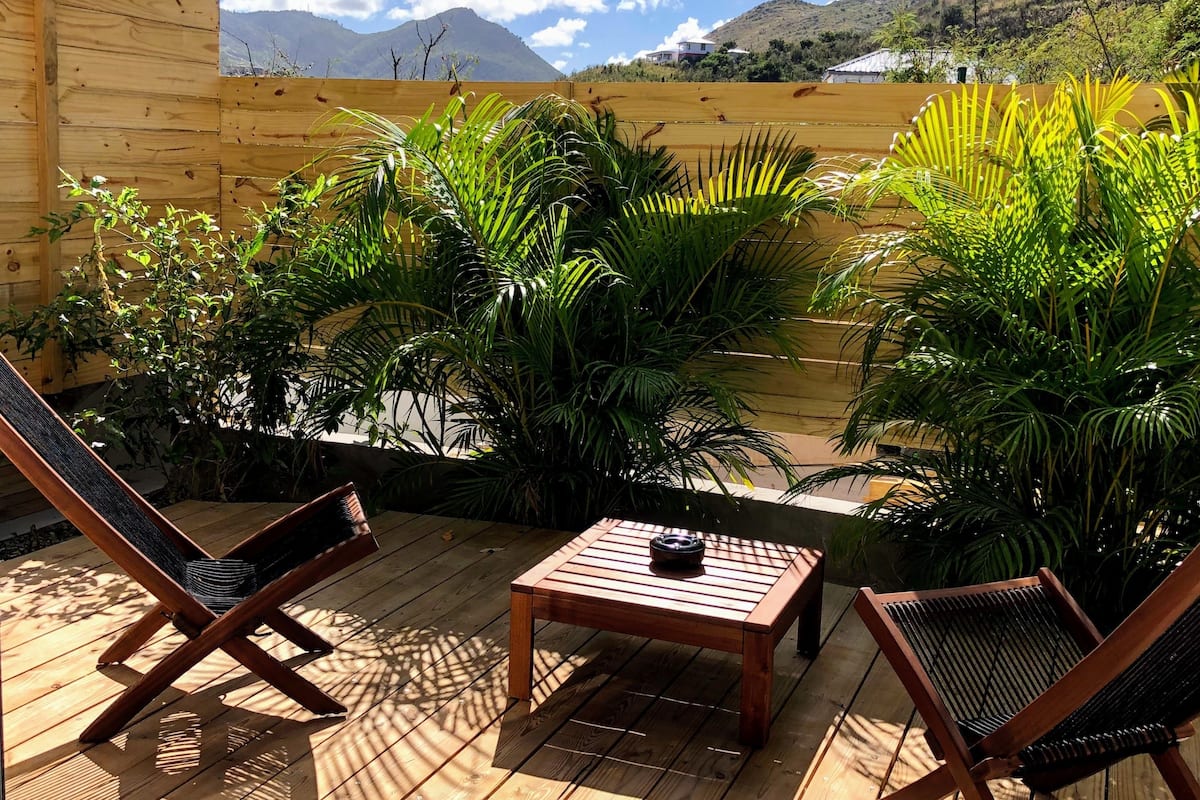 A wooden deck with two lounge chairs and a table, surrounded by lush tropical plants and a view of a mountainous landscape.