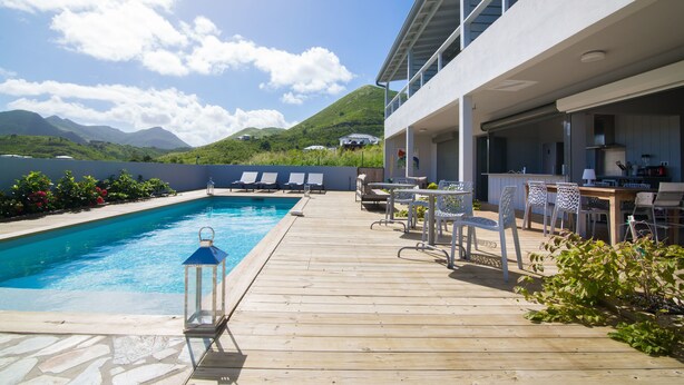 A wooden deck with a swimming pool, lounge chairs, and a bar area overlooking a mountainous landscape. - Villa Jwi Lavi Boutique Hotel (Cul de Sac)