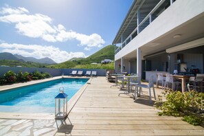 A wooden deck with a swimming pool, lounge chairs, and a bar area overlooking a mountainous landscape. - Villa Jwi Lavi Boutique Hotel (Cul de Sac)