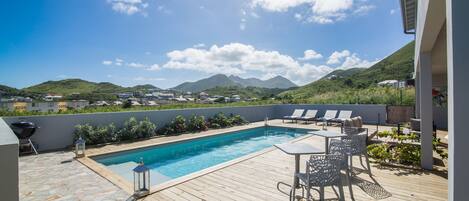 A rooftop pool area with a wooden deck, lounge chairs, and a view of mountains in the background.