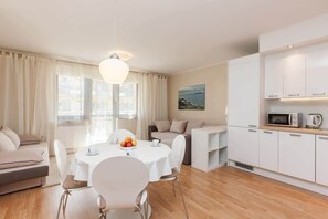 A modern, well-lit living area with a round dining table, white chairs, a sofa, and a kitchen area with white cabinets and appliances.