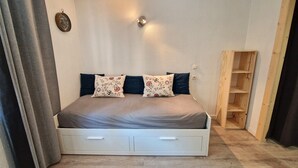 A bedroom with a bed, two pillows, a bedside table, a wall clock, and a wooden shelf unit. - Charming Studio for 4 in Resort Center, Steps from Slopes (Valfréjus)