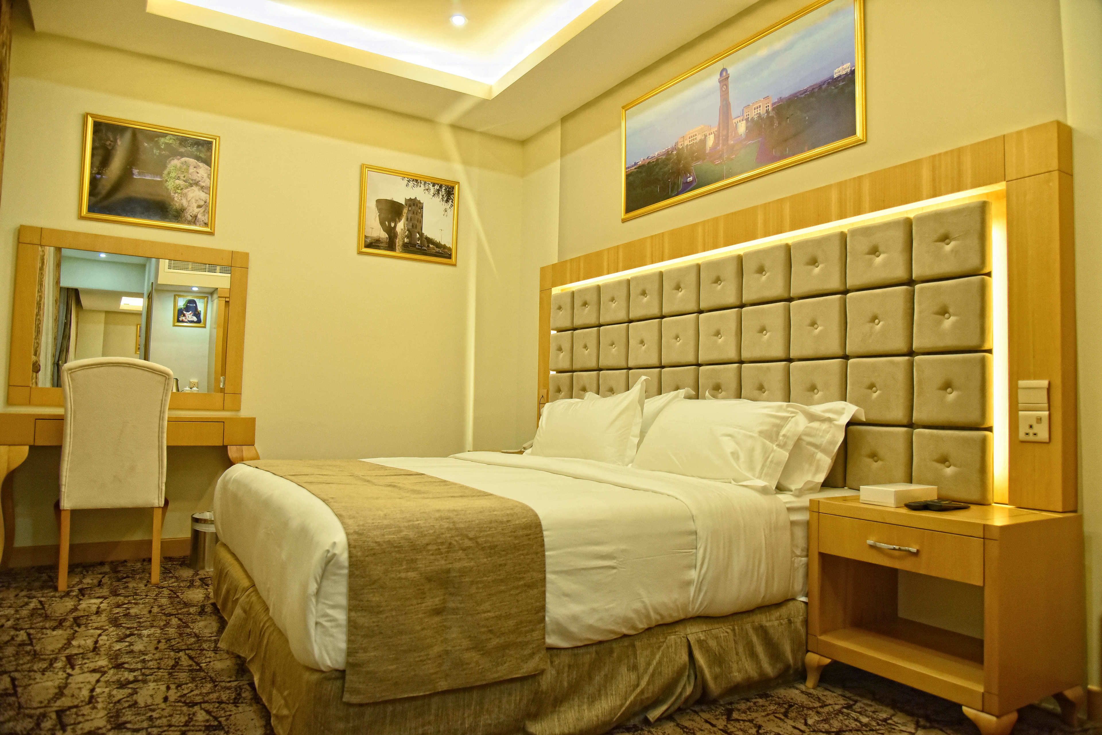 A hotel room with a large bed, a bedside table, a desk with a chair, a mirror, and two framed pictures on the wall.
