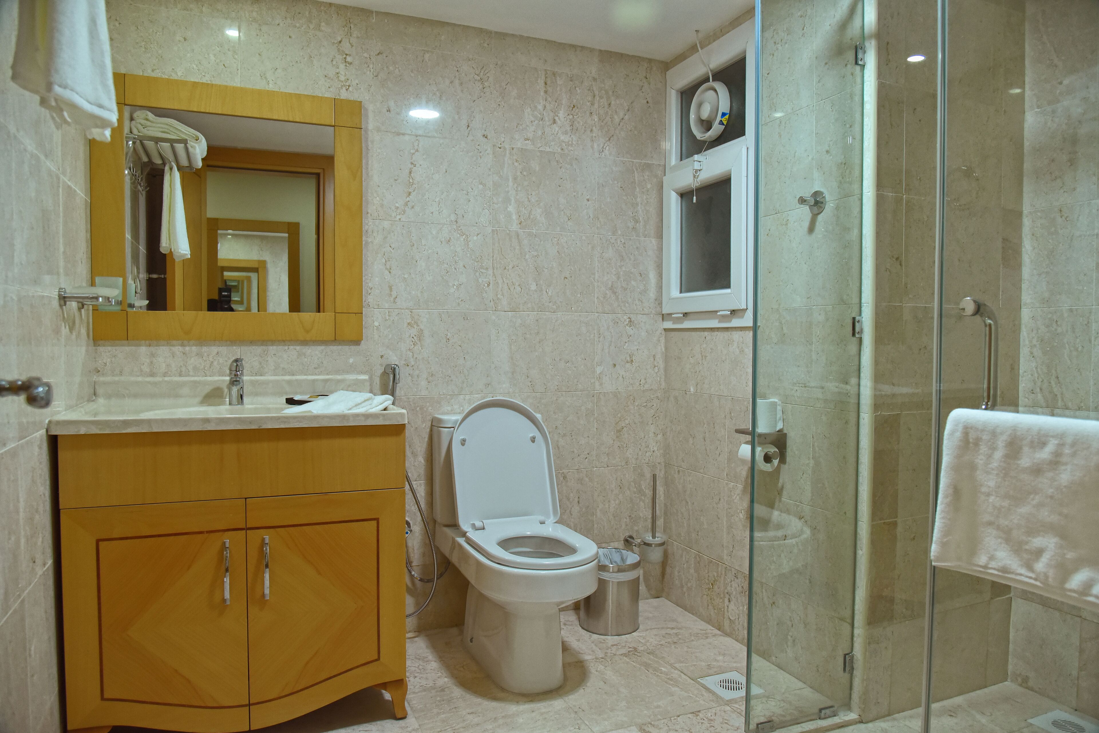 A modern bathroom with a marble floor, a wooden vanity, a white toilet, a glass shower enclosure, and a mirror with a towel hanging on it.