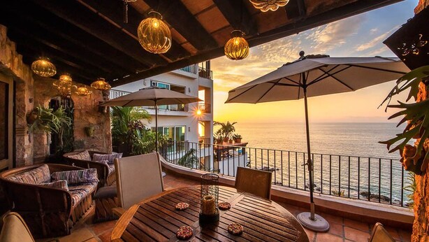 A terrace with wicker furniture, a wooden table, and two umbrellas overlooking the sea at sunset. - 2 BR romantic Penthouse in Conchas Chinas with amazing views & beach access (Puerto Vallarta)