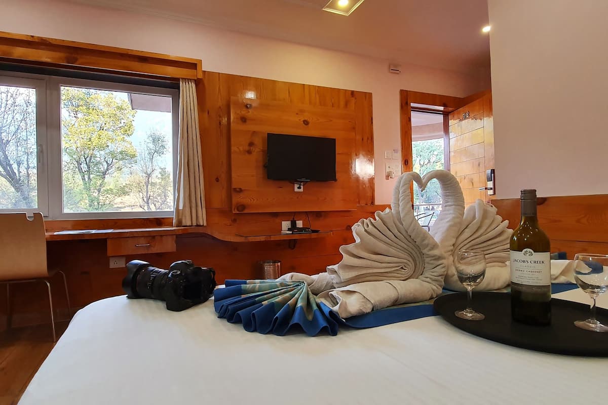 A room with wooden walls, a large window with curtains, a flat-screen TV mounted on the wall, a bed with a white comforter, a bottle of wine, and a glass on a black tray.