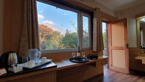 Habitación de hotel con un amplio ventanal que ofrece vistas a una montaña y árboles. Hay un escritorio de madera con una cafetera, tazas y una copa de vino.