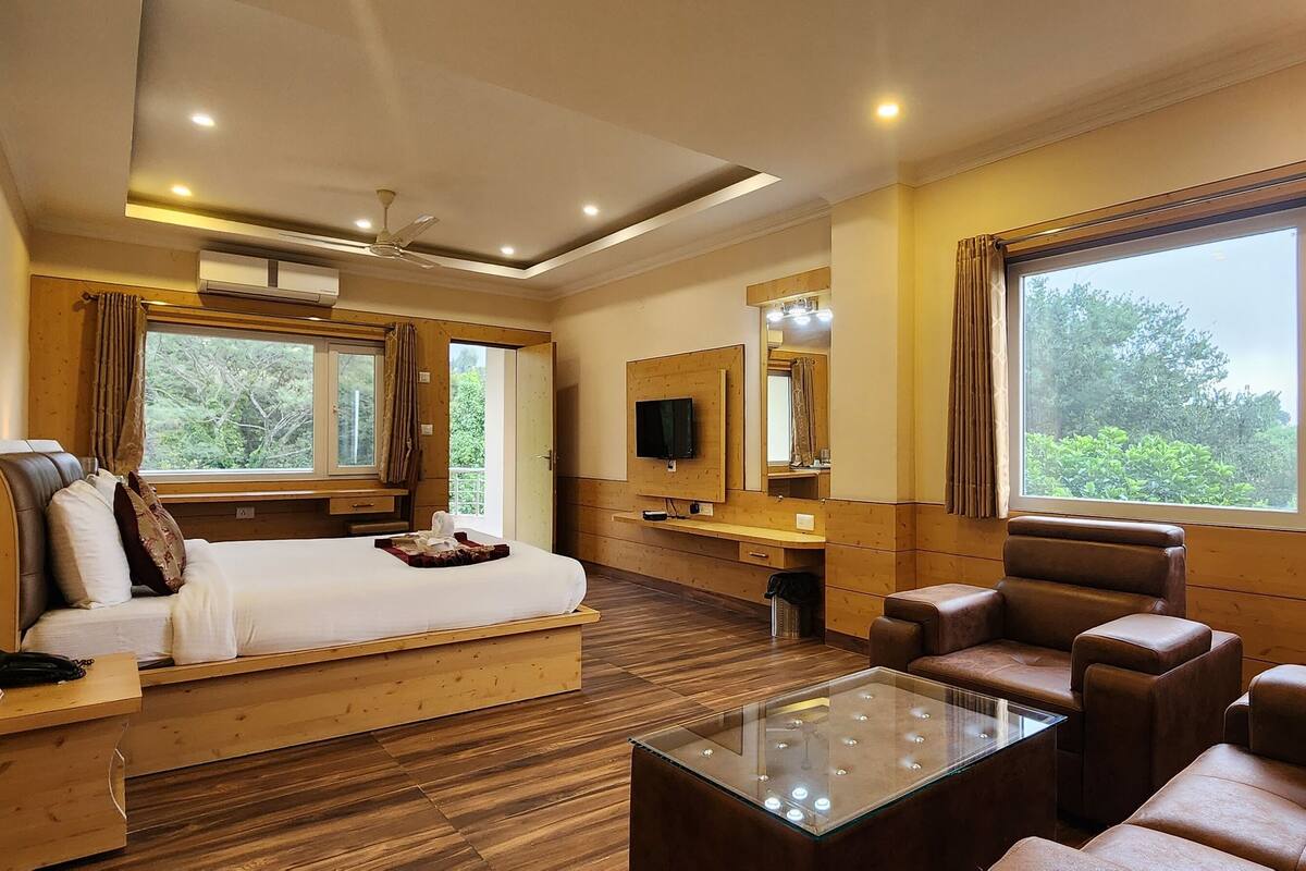 A hotel room with a bed, a sofa, a coffee table, a TV, and large windows with curtains.
