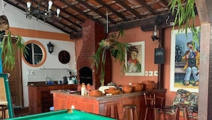 A pool table with a green felt surface, a bar area with stools, and a kitchen with a brick wall and hanging plants. - Aloha Hostel Cabo Frio (Cabo Frio)