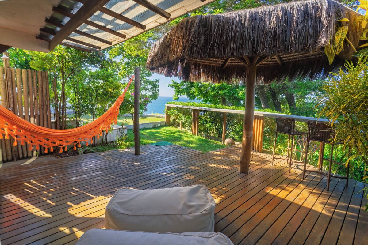 Une terrasse en bois avec un hamac, une structure à toit de chaume et une vue sur la mer.