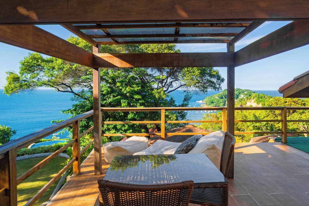 Un espace extérieur abrité, aménagé avec des places assises, offrant une vue sur la mer, doté de rampes en bois et d’une table avec des coussins.