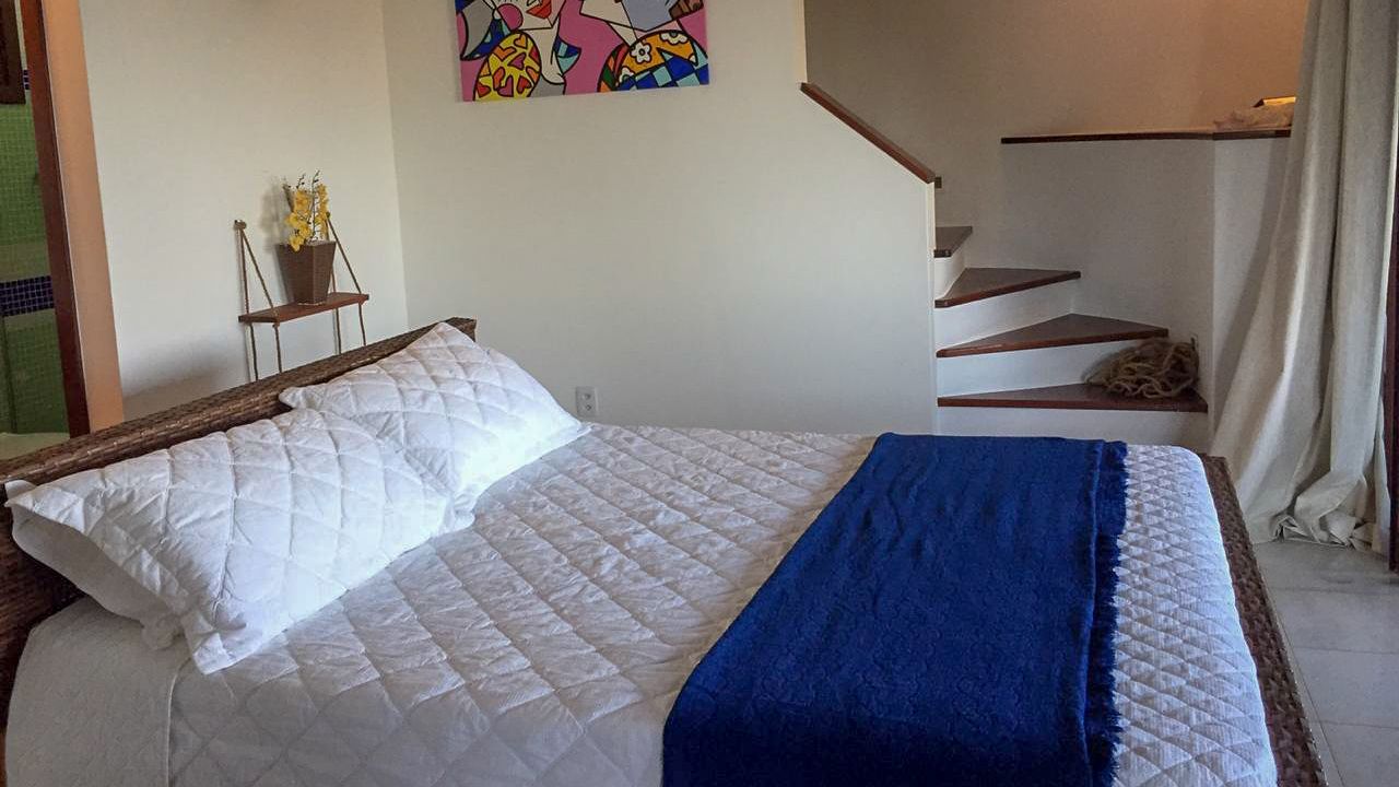 A bedroom with a bed, a blue blanket, a white quilted bedspread, a colorful abstract painting on the wall, and a staircase with a bag on the landing.