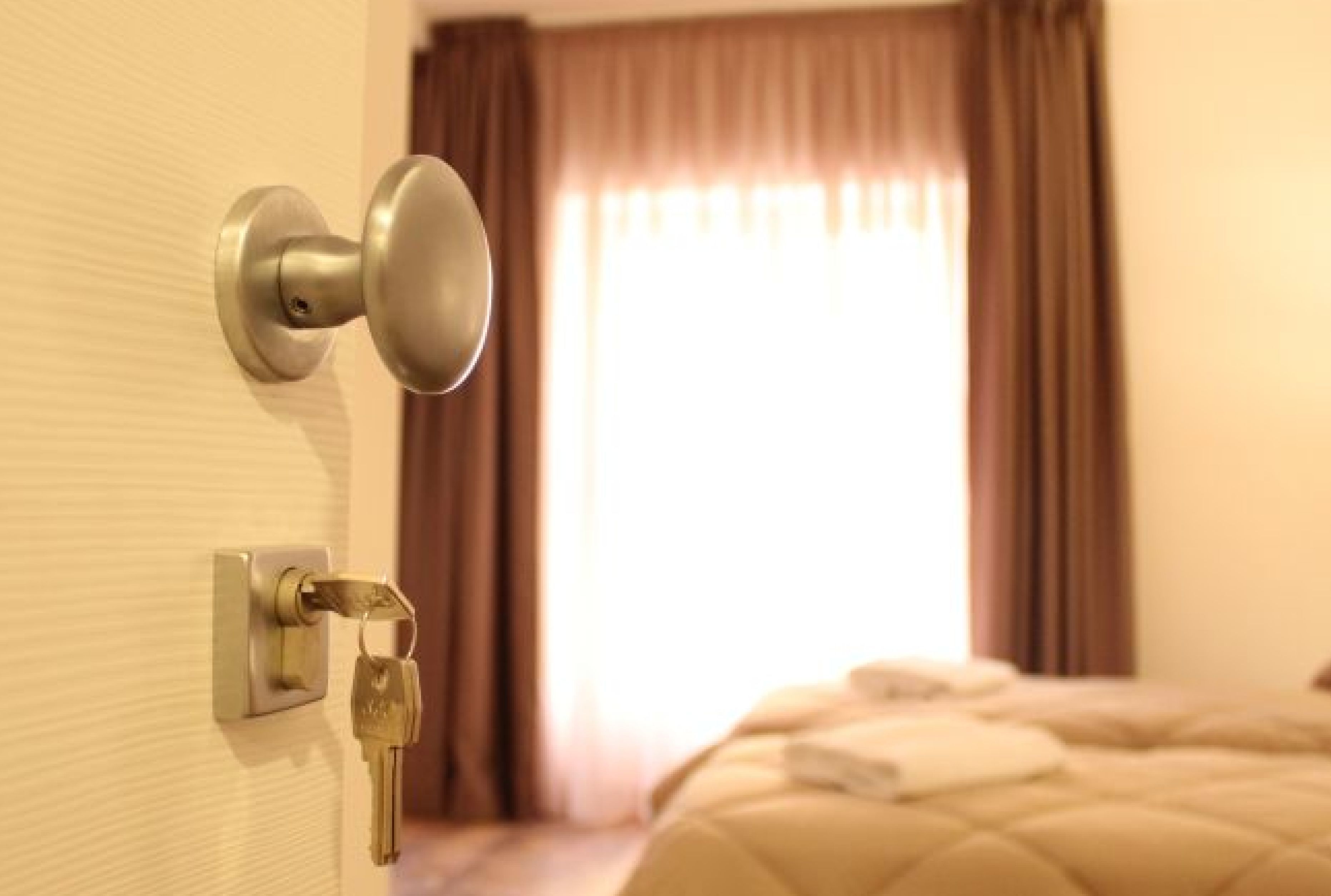 A hotel room with a door, a key in the lock, a bed with a quilted cover, and a window with curtains.
