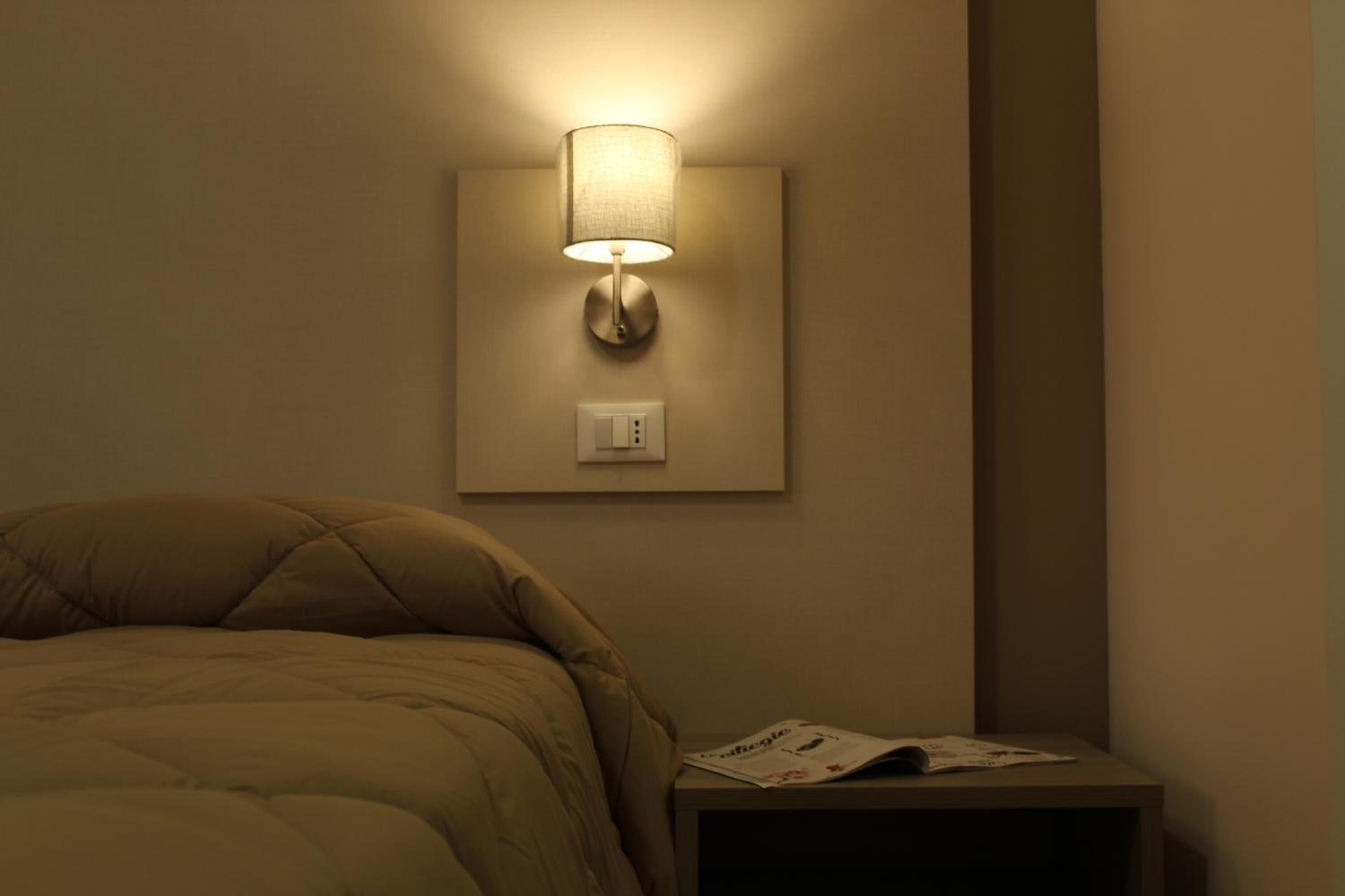 A bedroom with a bed, a nightstand, a wall-mounted lamp, and a power outlet.