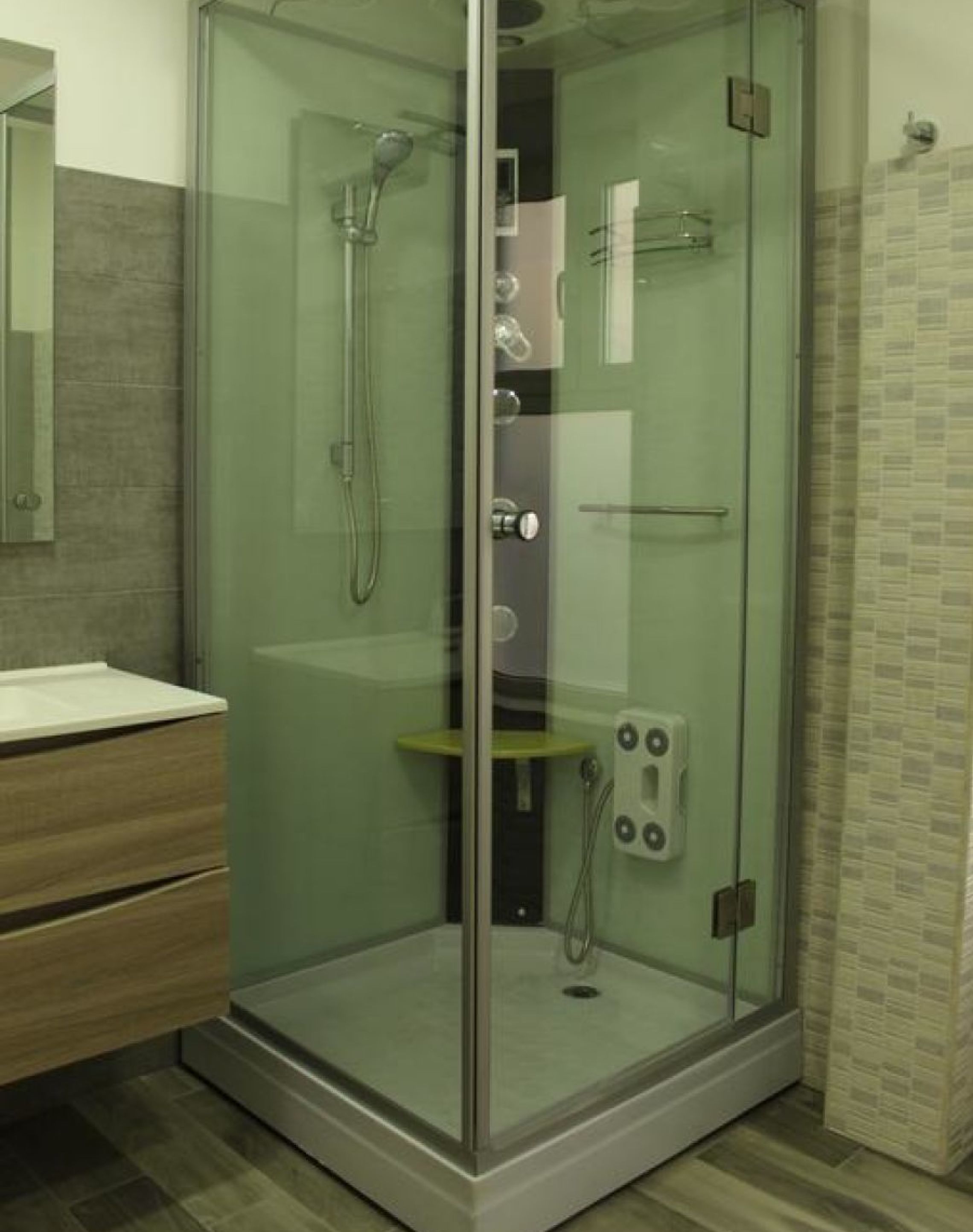 A glass-enclosed shower stall with a handheld showerhead, a wall-mounted faucet, and a small shelf.