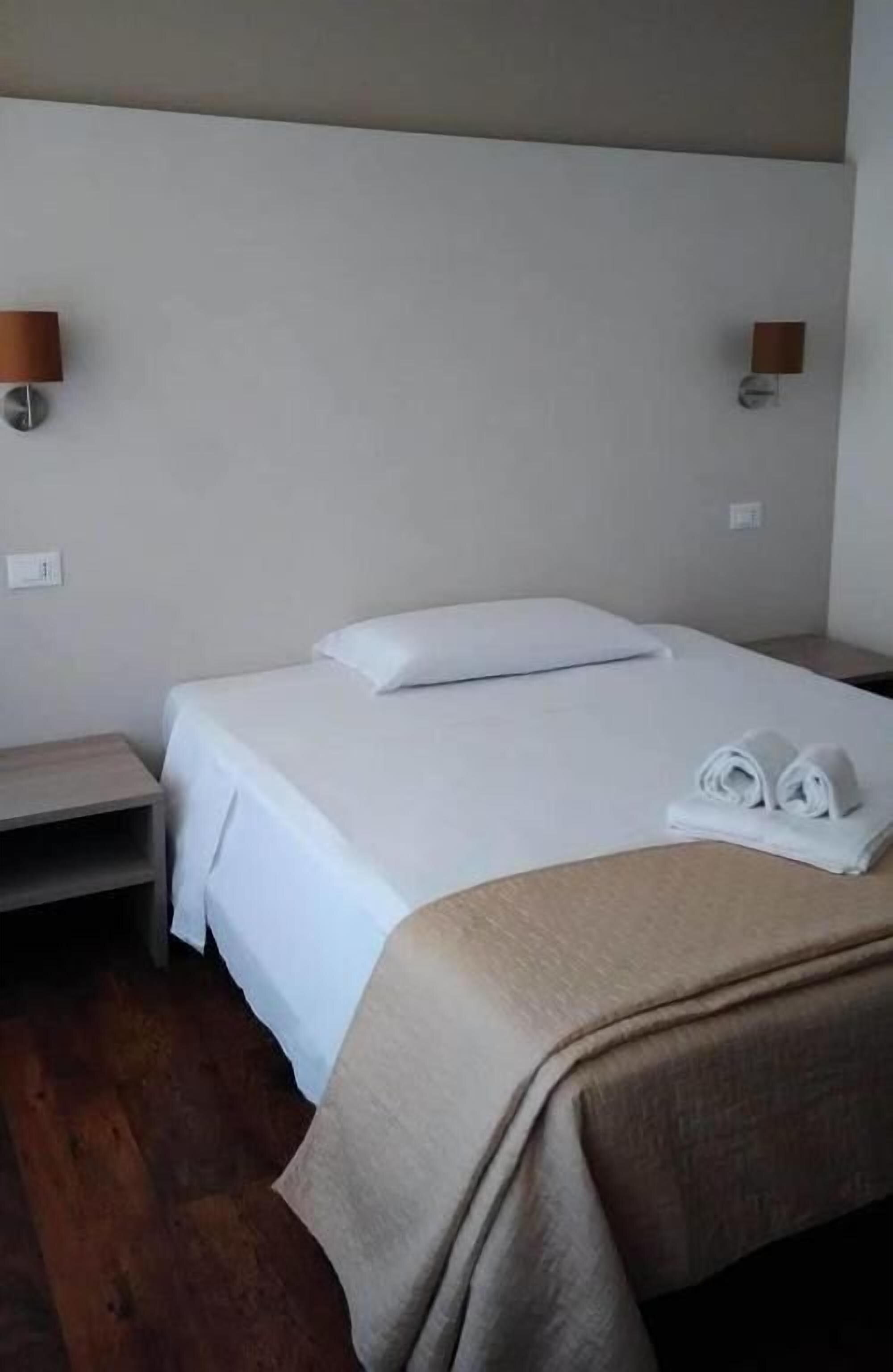 Una cama individual con ropa de cama blanca, una mesita de noche y dos lámparas fijadas a la pared.