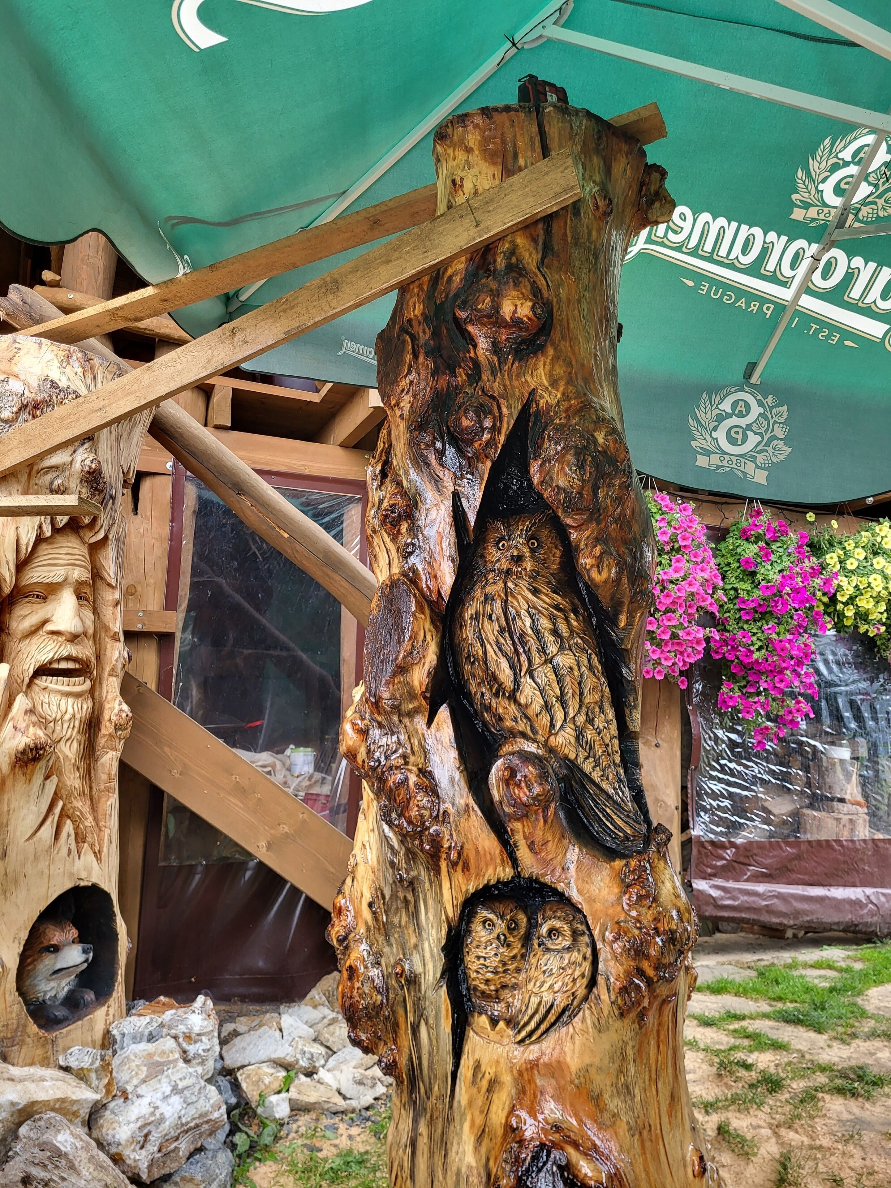 A wooden sculpture with carved owls and a fox, set against a backdrop of a green awning and pink flowers.