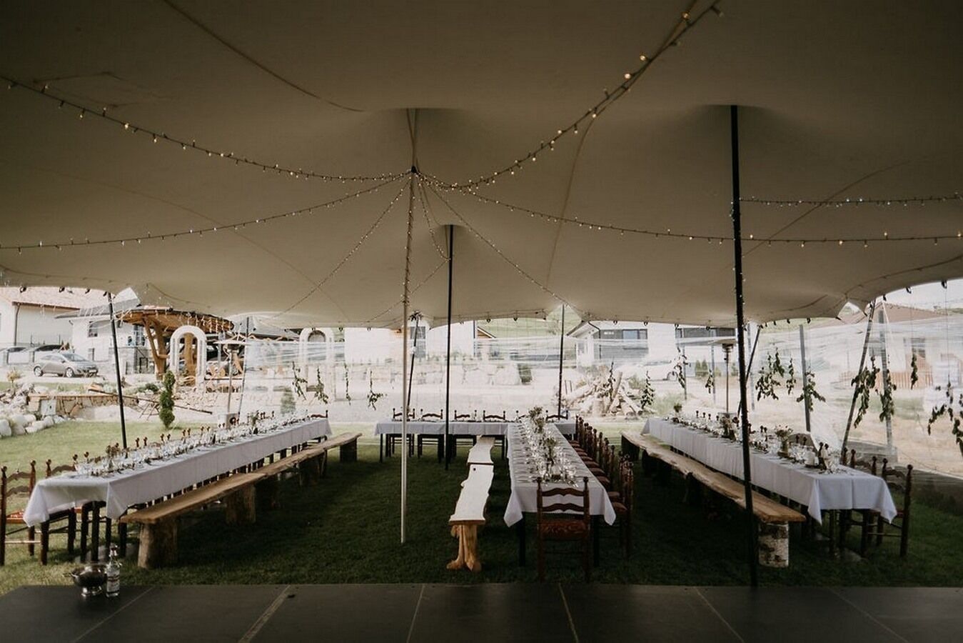 An outdoor tented dining area with tables set for a meal, surrounded by a garden and a view of a building in the background.
