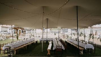 An outdoor tented dining area with tables set for a meal, surrounded by a garden and a view of a building in the background.