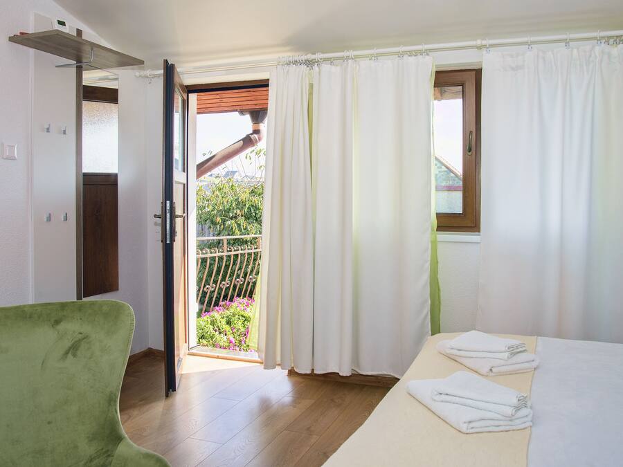 A room with a bed, a green chair, a balcony with a view of greenery, and white curtains.