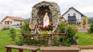 A stone structure with a statue, surrounded by flowers and benches in a grassy area with houses in the background. - VeLa (Malý Slavkov)