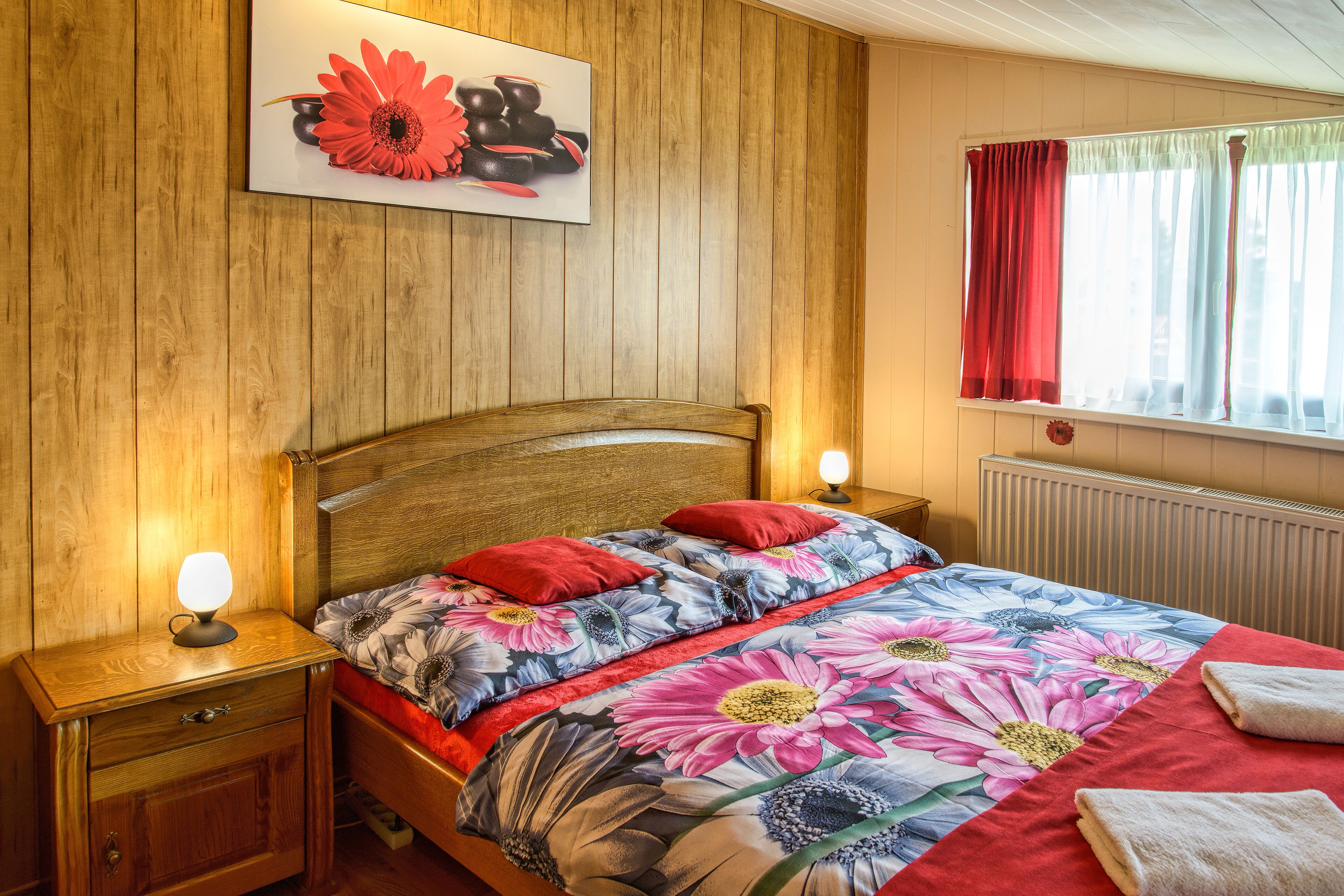 A bedroom with a wooden headboard, a floral bedspread, red pillows, and a bedside table with a lamp.