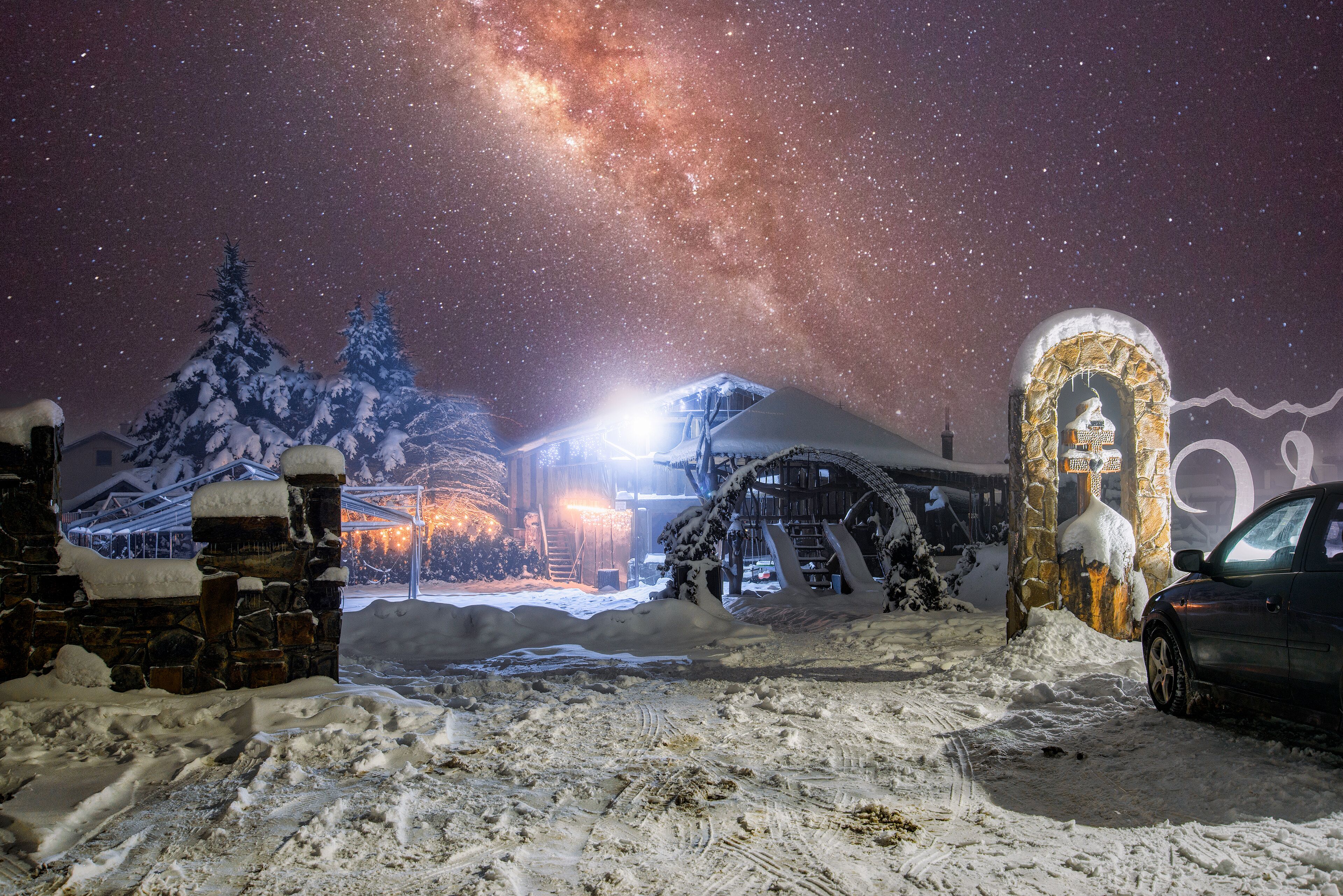 A snowy landscape with a stone archway, a building with a snow-covered roof, and a car parked nearby.