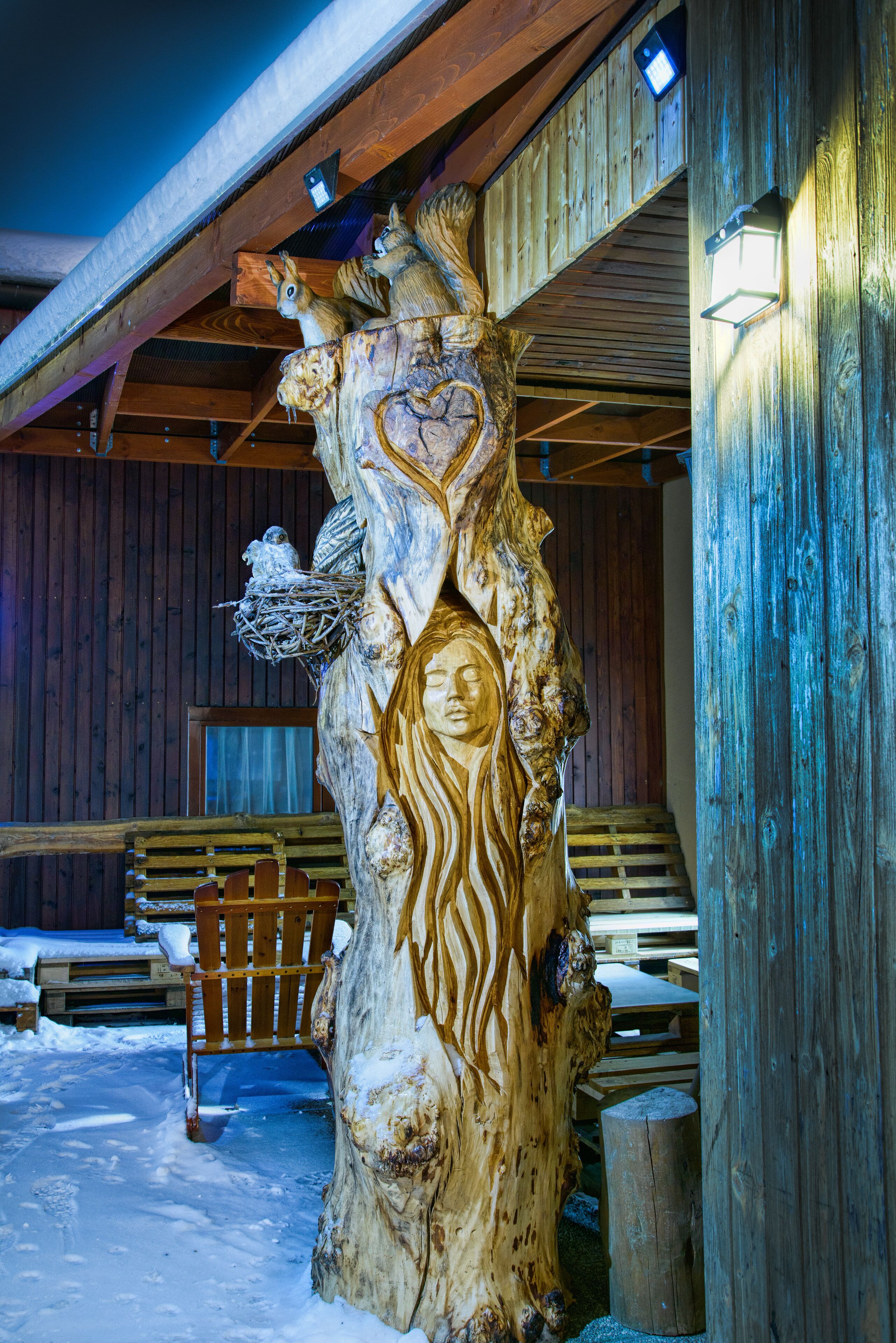 A wooden sculpture with carved figures and a bench in a snowy outdoor area.
