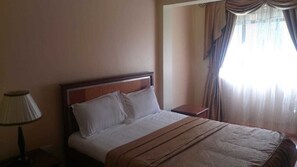 A hotel room with a bed, a nightstand, a lamp, and a window with curtains. - Pinnacle Court Apartments (Nairobi)