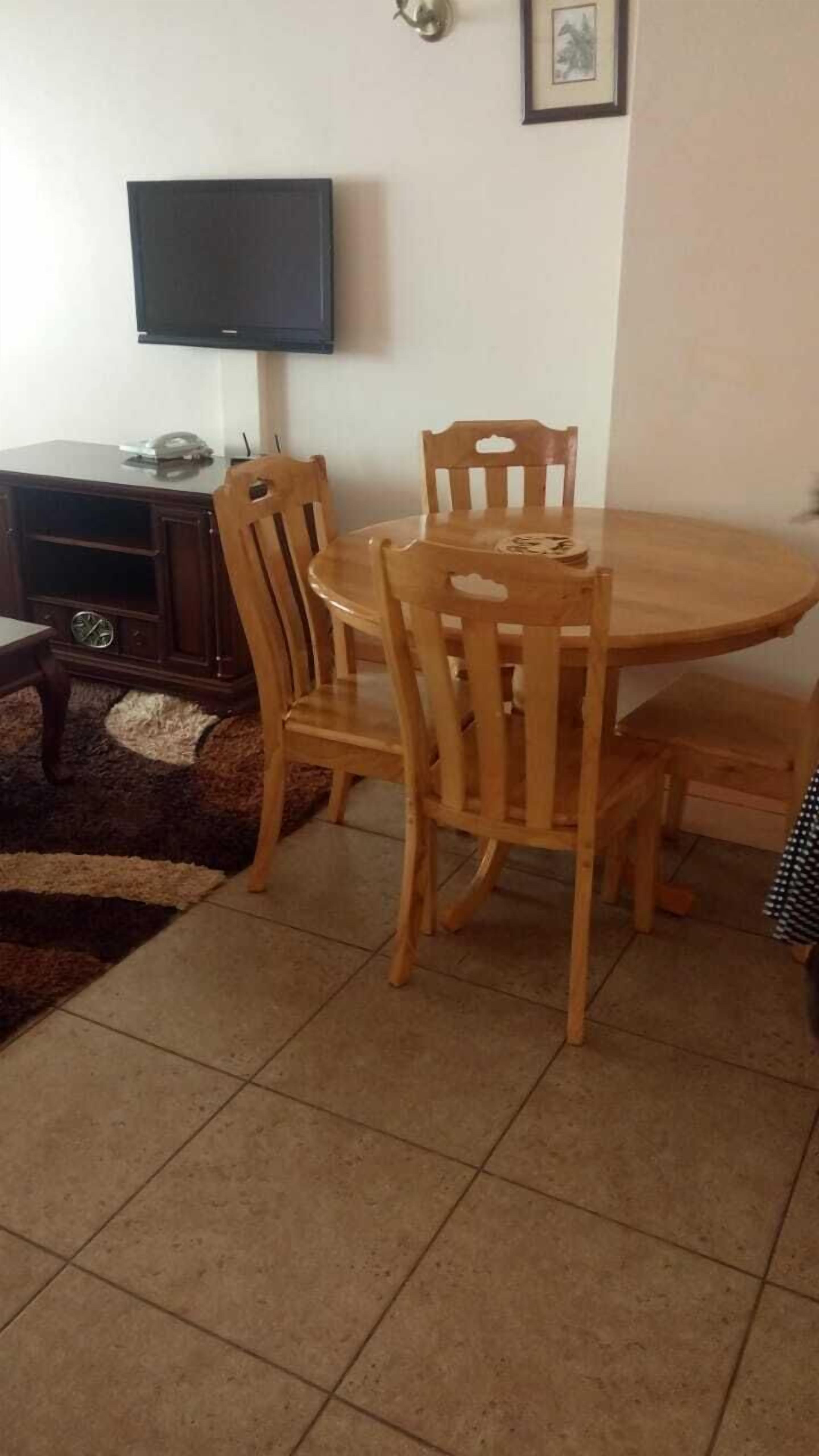 Uma sala de jantar com mesa e cadeiras de madeira, uma televisão na parede e um pequeno armário com relógio.