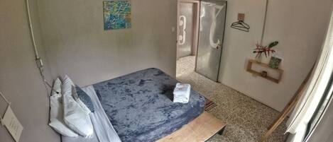 A bedroom with a bed, a bedside table, a hanging light, a door, and a small shelf with a plant.