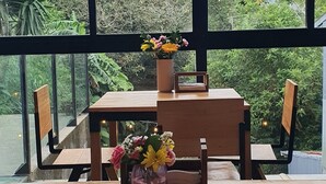 Et træbord med stole og en vase med blomster, foran et stort vindue med udsigt over en frodig have.