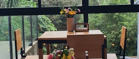 A wooden table with chairs and a vase of flowers, set against a large window overlooking a lush garden.