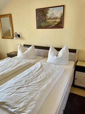 A double bed with white linens and pillows, a painting of a landscape, a mirror, and a nightstand with a lamp.