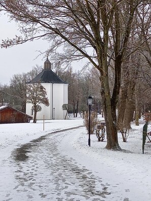 A snow-covered path leading to a white building with a dark roof, surrounded by bare trees and a small red structure.
